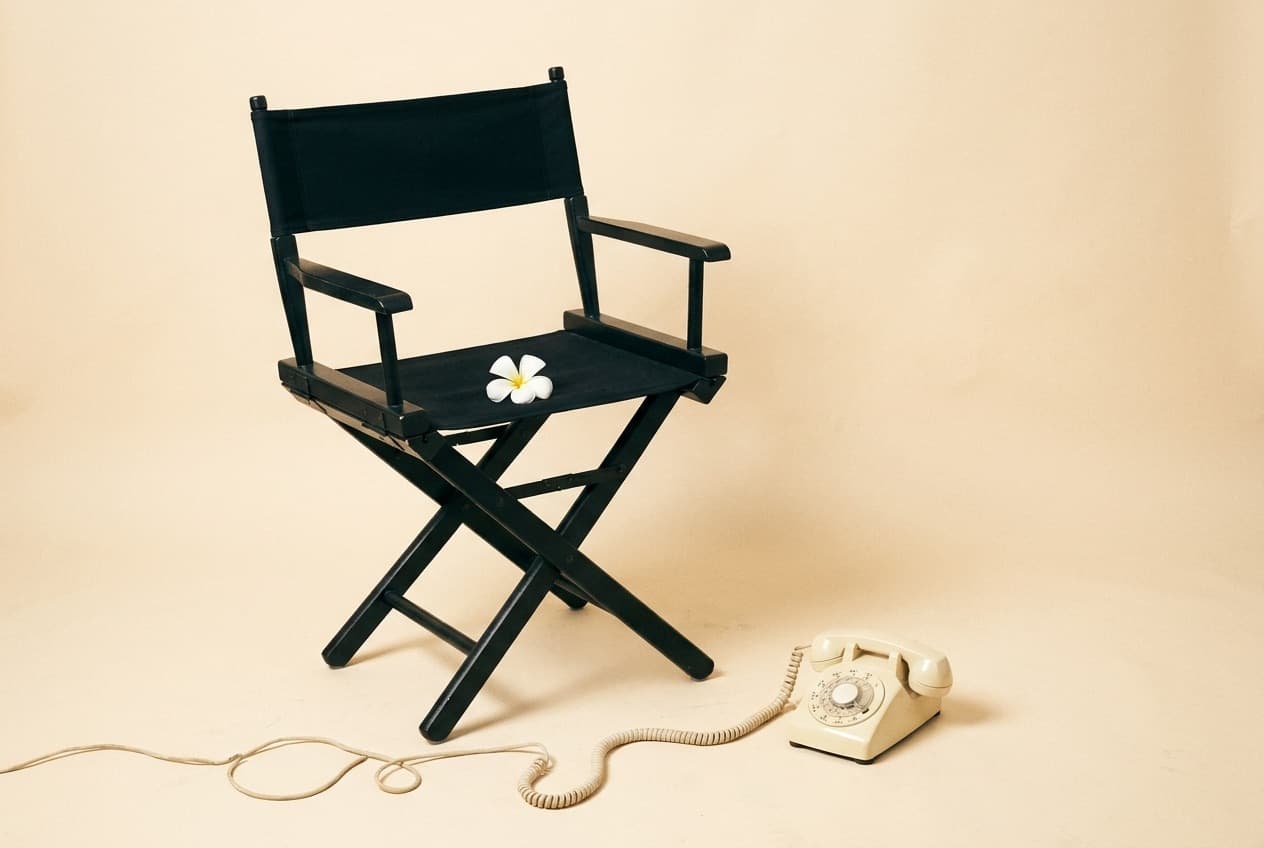 An empty black director's chair on a warm cream backdrop, a white plumeria resting on the seat and a cream rotary telephone on the floor beside it.